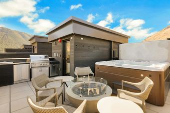 A spacious rooftop terrace with a hot tub, BBQ grill, fire pit, and comfortable seating, all set against a backdrop of mountains under a blue sky.