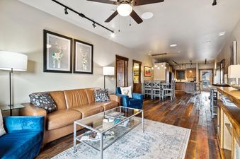 A spacious open-concept living room and dining area in Baker Loft 302 features a brown leather sofa, blue armchairs, a glass coffee table, and a dining table with chairs, all set on dark wood floors.