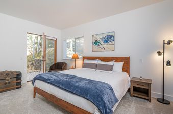 A bright bedroom with a wooden bed, white and blue bedding, a skiing artwork, and access to an outdoor patio.