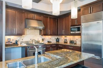 The kitchen in Lumiere One Bedroom Standard Residence has stainless steel appliances, granite countertops, a double sink, and a view of the living area.