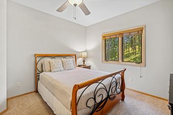 A cozy bedroom with a bed featuring a wooden and wrought iron frame, white bedding, and a nightstand with a lamp, offering a view of green trees through a window.