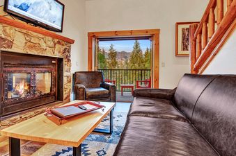 A cozy living room at the Mountain Lodge 2 Bedroom Suite features a stone fireplace, leather furniture, and a balcony with mountain views.