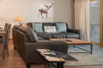 A cozy living area features a dark brown sectional sofa with light blue pillows and a decorative 'Gone Skiing' pillow, a rustic wooden coffee table with a remote control, and a modern side table with an abstract design.