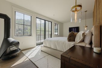 A bright and airy master bedroom with a king-size bed, a modern black fireplace, and large windows offering a view of a forested mountain.