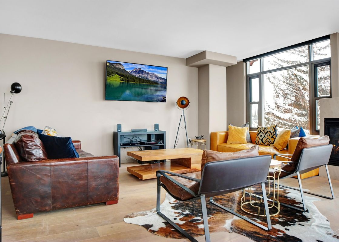 A cozy and modern living room at Lowell 301 features leather sofas, a flat-screen TV, a rustic coffee table, and a fireplace, with large windows overlooking a snowy landscape.