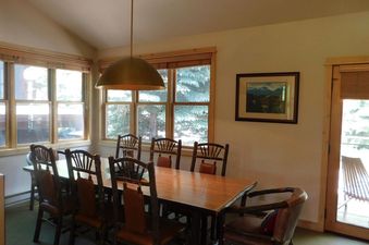 A spacious dining area at Moose Creek 35 features a large wooden table with rustic chairs, illuminated by a brass dome pendant light and natural light from multiple windows showcasing evergreen trees.