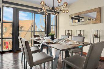 A modern dining table set with white dishes and clear glasses in a dining room with large windows overlooking buildings and a blue sky.