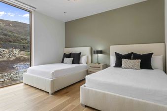 A serene guest room at Founders Place 1504 features two twin beds with white linens, dark pillows, and a window offering a view of the natural landscape.