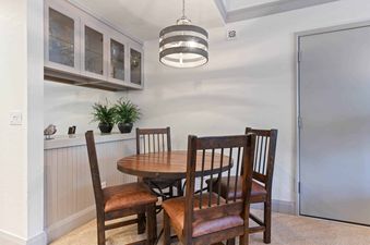 A cozy dining area featuring a round wooden table, four chairs with leather seats, a drum-style chandelier, and a built-in cabinet with glass doors displaying glassware.
