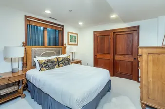 A cozy bedroom featuring a bed with a decorative headboard, two nightstands with lamps, a window with wooden blinds, and wooden closet doors, all enhancing the inviting ambiance of the South Pacific Townhouse.