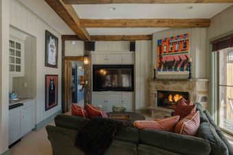 A cozy lounge in Elk Lake Lodge with a large television, a stone fireplace, and rustic wooden beams overhead.