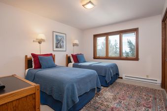A cozy bedroom in Teewinot D4 with two twin beds dressed in blue bedding, a patterned area rug, and a large wooden-framed window looking out onto trees.