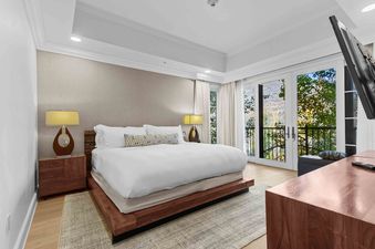 A white king-sized bed with white linens and a geometric patterned throw pillow sits in the center of a bedroom with hardwood floors.