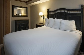 A cozy bedroom at Summit Suite Beaver Run Resort featuring a bed with white bedding, a dark wooden headboard, a dresser with a mirror, and a nightstand with a wall-mounted lamp.