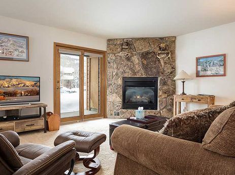 A cozy living room in Pines Condominiums Two Bedroom with a stone fireplace, comfortable seating, and a flat-screen TV, leading to a snowy patio.
