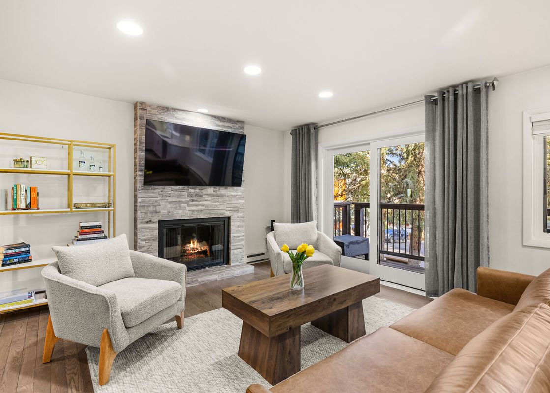 A modern living room with a tan leather sofa, two armchairs, a wooden coffee table, a gold and white bookshelf, a stone fireplace, and a large television.