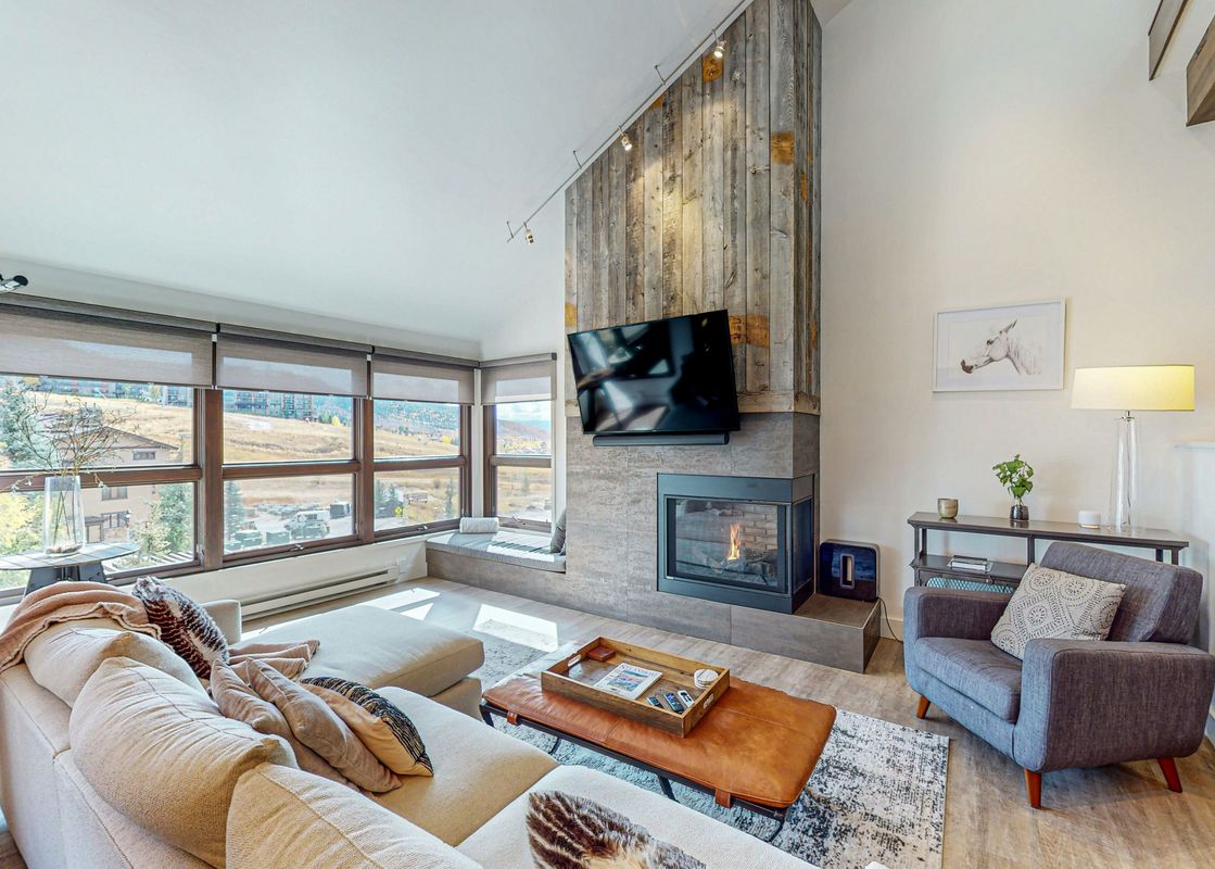 A living room with a fireplace, large windows with a view of the mountains, a couch, a coffee table, and a chair.