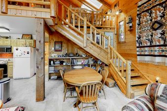 The dining area of Atkins Cabin features a wooden table and chairs, a bookcase filled with books and decor, and a view of the wooden staircase leading to the loft.