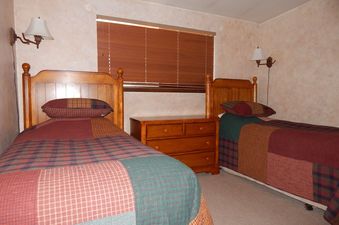 The two wooden twin beds are adorned with colorful plaid bedding, set in a cozy bedroom with light-colored walls and a window featuring wooden blinds.