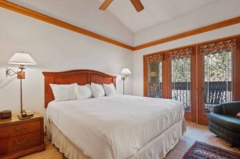A spacious bedroom featuring a king-sized bed, elegant wooden furniture, and double doors with leaded glass opening to a balcony with forest views.