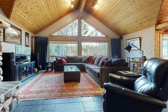 A cozy living room with an L-shaped sofa, a large area rug, and windows offering views of nature.
