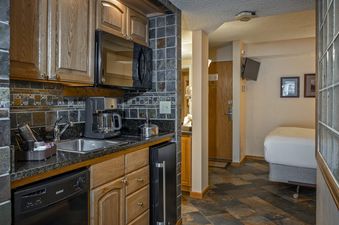 A fully equipped kitchenette with wooden cabinets, granite countertops, and modern appliances is shown next to a living area with a bed and television, all on a tiled floor.