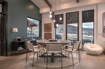 A modern dining room with a round wooden table set for a meal, illuminated by unique pendant lights, featuring large windows with a view of a snowy mountain landscape.