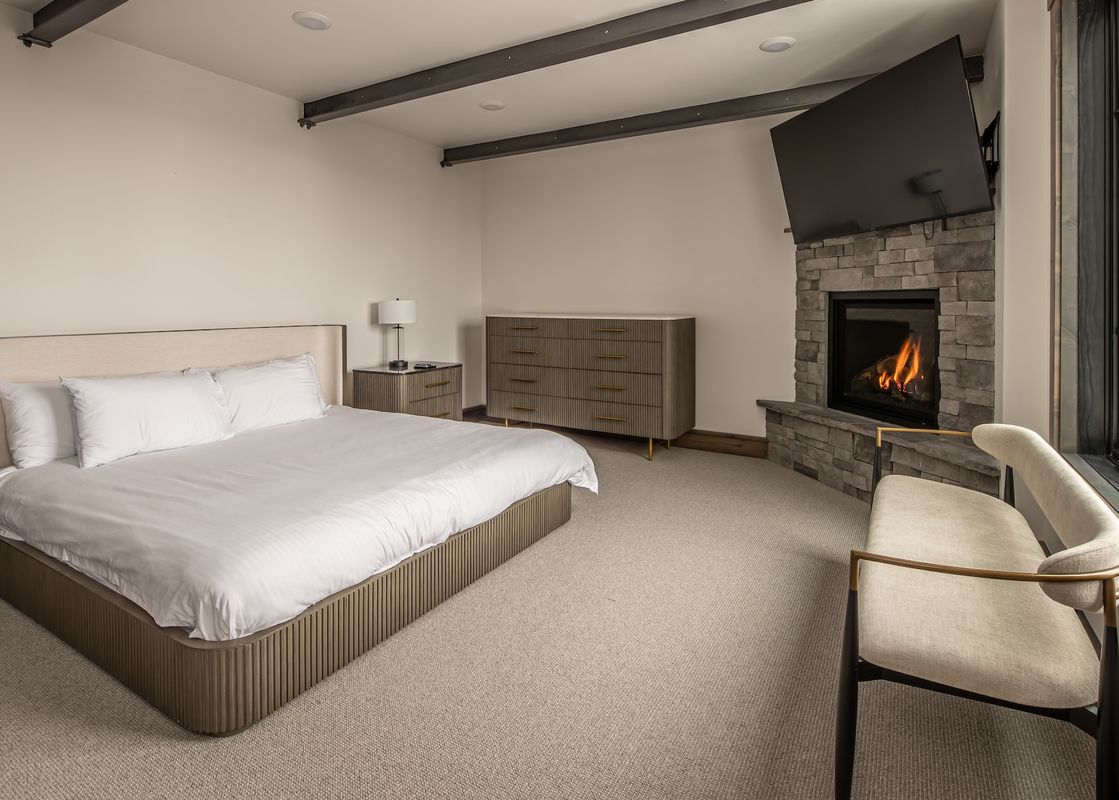 A modern master bedroom featuring a king-sized bed, a stone fireplace with a TV above, and a plush bench by a large window.