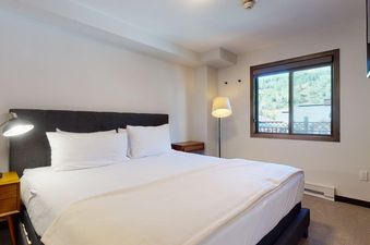 A cozy bedroom in Landmark 704 features a queen-sized bed with white bedding, wooden nightstands with lamps, and a window offering a view of green mountains.