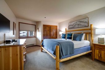 A cozy bedroom with a rustic log-frame king-size bed, blue bedding, a television on a wooden dresser, and a window with a pleated blind.