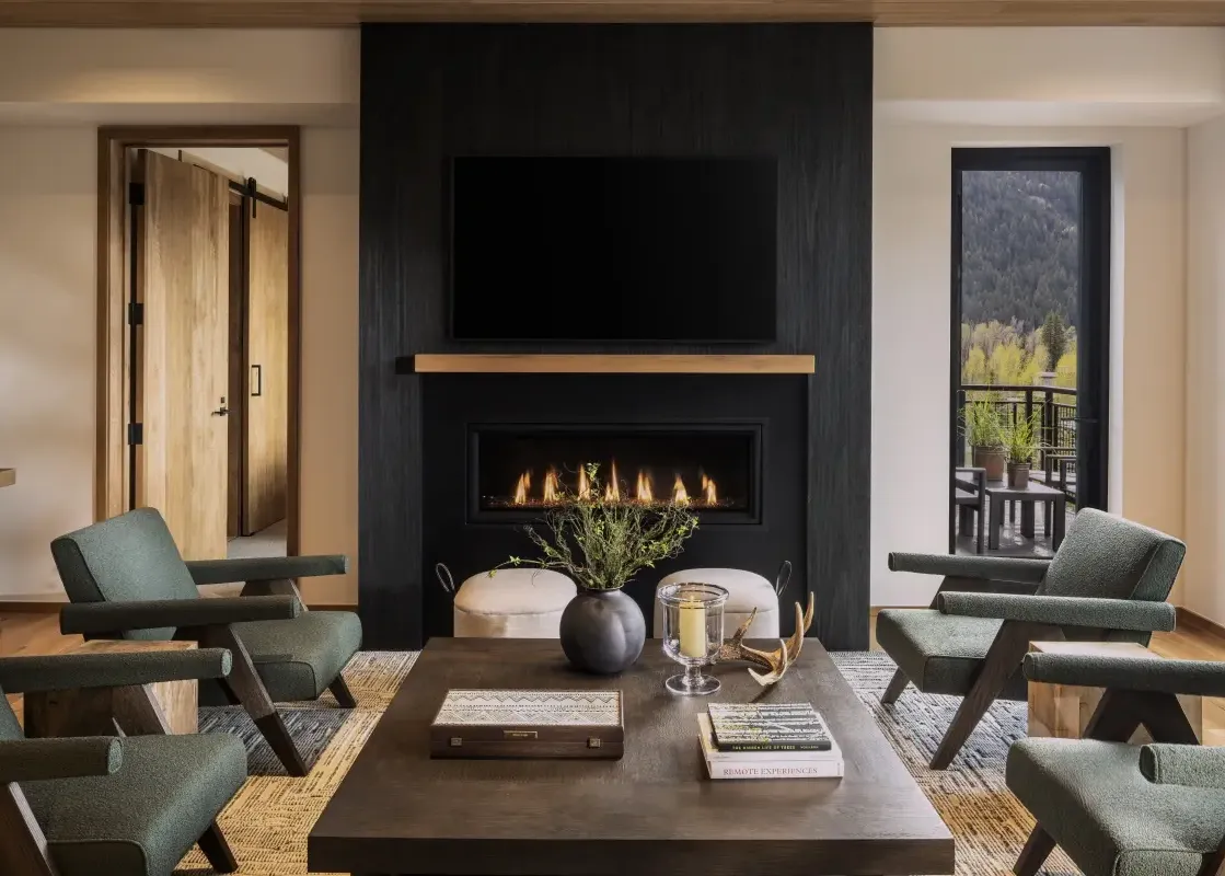 A luxurious living room in The Sylvan Lodge Two Bedroom Suite features a black fireplace, a television, four green armchairs, and a large coffee table, with a stunning mountain view from the window.