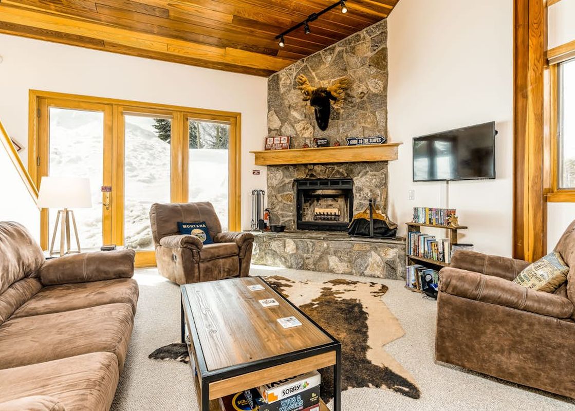 A cozy living room in Big Horn 65 with a stone fireplace, moose head trophy, brown sofa and armchair, coffee table, and a large window looking out onto a snowy landscape.