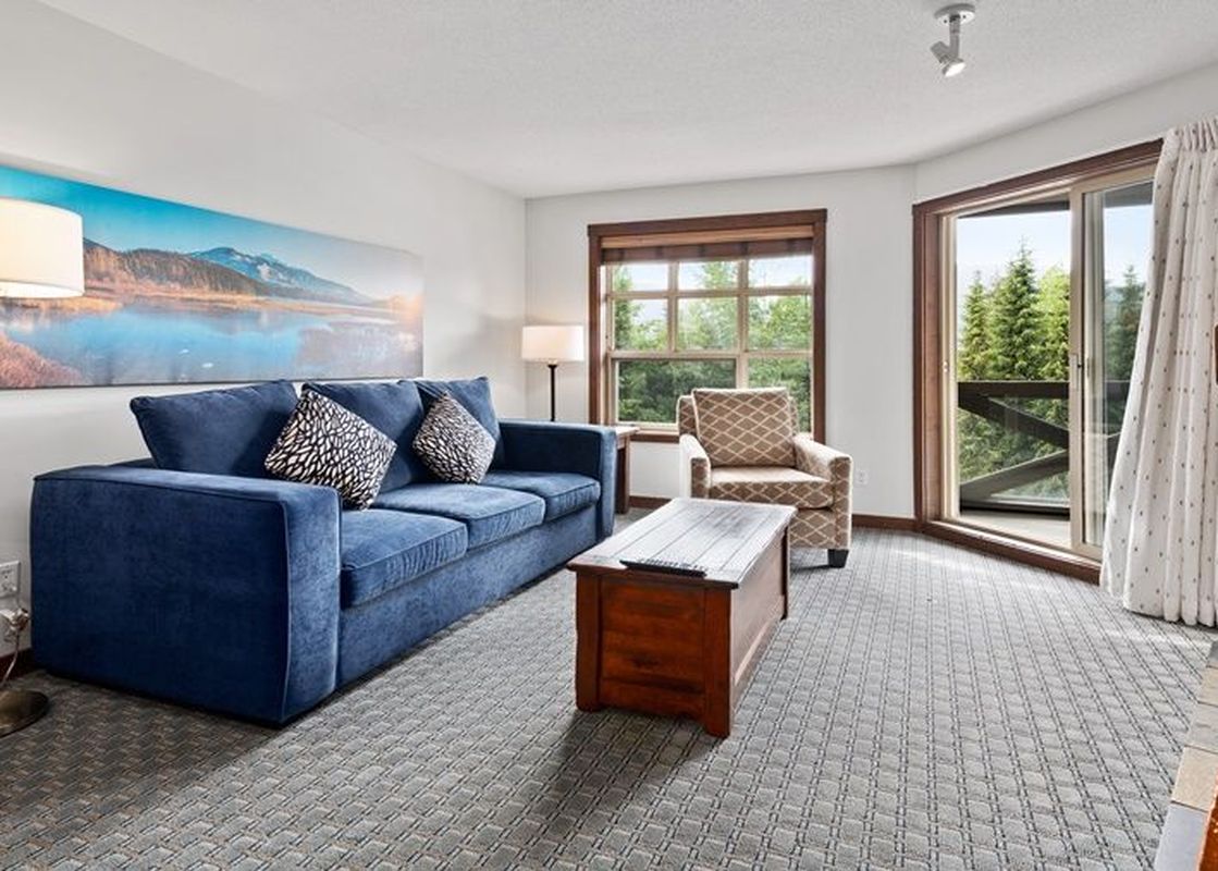 The living area of the Blackcomb Springs Two Bedroom Slopeside Suite features a plush blue sofa and a natural wood coffee table, set against a backdrop of a large landscape painting.