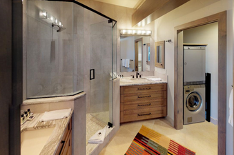 A modern bathroom with a large shower, a double vanity, and a washer and dryer in the adjacent room.