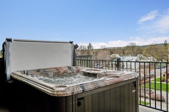 A bubbling hot tub on a deck with an open cover, offering views of a town and hills under a blue sky.