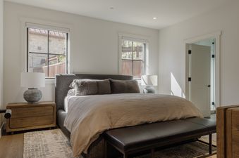 A cozy bedroom with a dark gray bed, two windows, nightstands with lamps, and a leather bench.