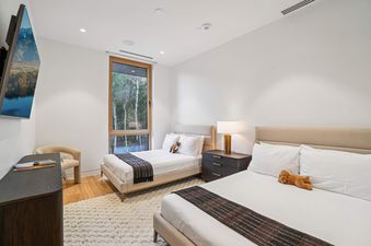 A modern guest bedroom with two queen beds, a large window revealing a wooded view, and a wall-mounted television.