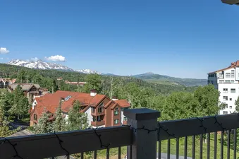 A view from a balcony with a wooden railing overlooking a forest and mountains in the distance.