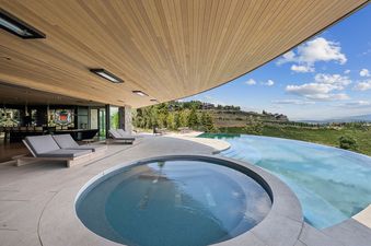 An expansive outdoor patio with a large swimming pool and hot tub, offering panoramic views of a lush green hillside and distant mountains under a blue sky.