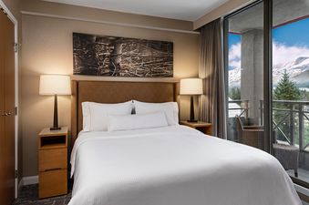 A cozy bedroom with a large bed, two nightstands, and lamps. A balcony offers a scenic mountain view.