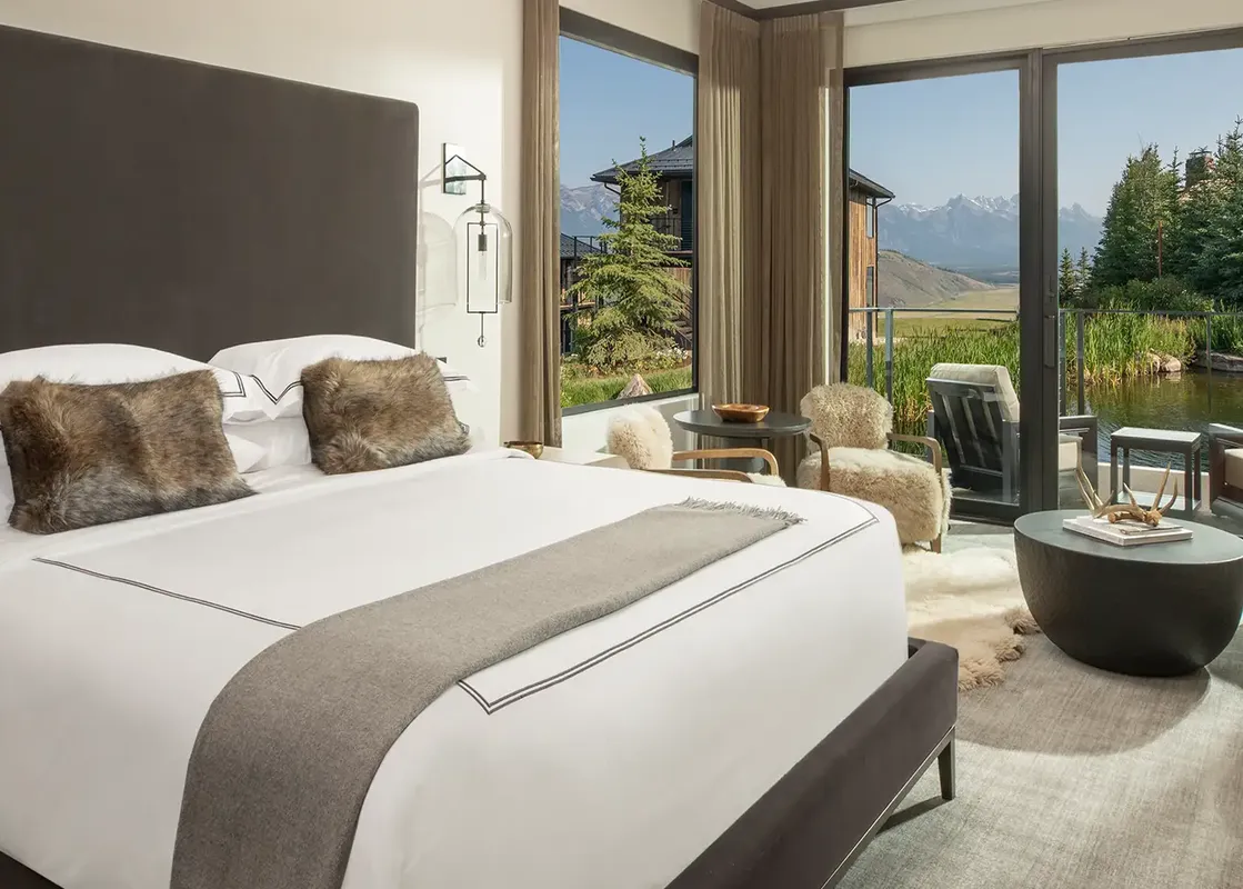 A luxurious bedroom, the Grand Reflection Suite, features a king-sized bed, a cozy seating area, a fireplace, and a large window overlooking a serene pond and majestic mountains.