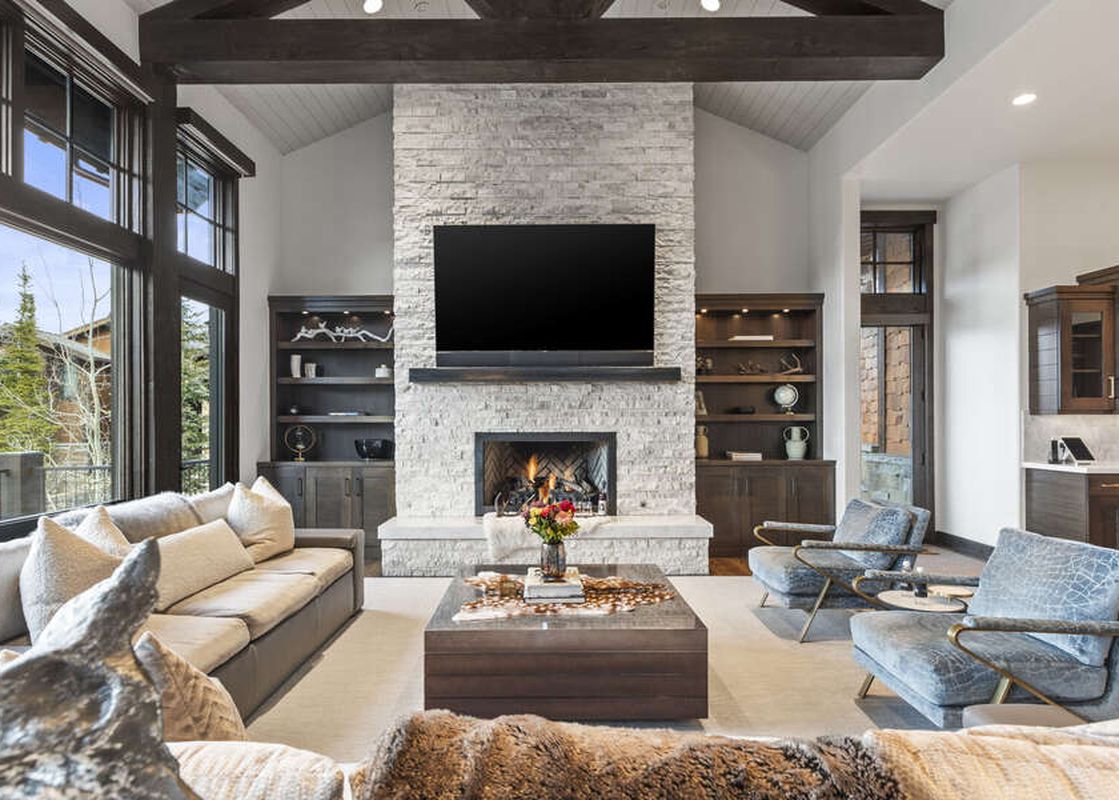 The spacious living room at Ruby Run features a large stone fireplace with a TV mounted above, flanked by built-in shelves, and comfortable seating arranged around a coffee table, with expansive windows offering outdoor views.