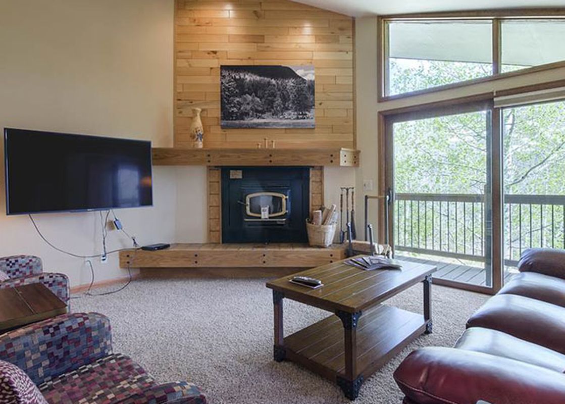 A cozy living room in Ranch at Steamboat RA604 with a brown leather sofa, patterned armchairs, a flat-screen TV, a fireplace, and a balcony with a view of green trees.