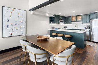 A modern dining area with a large wooden table and chairs, adjacent to an open-plan kitchen with dark green cabinetry and a white countertop.
