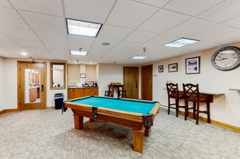 A spacious recreation room featuring a vibrant teal-felt pool table, bar-height seating, and a coffee station, all under well-lit ceilings.