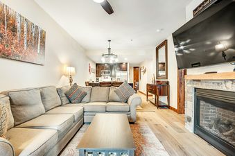 A cozy living room at BlueSky Breckenridge 407 featuring a large sectional sofa, a coffee table, a fireplace, and a flat-screen TV, seamlessly connected to a modern kitchen and dining area.