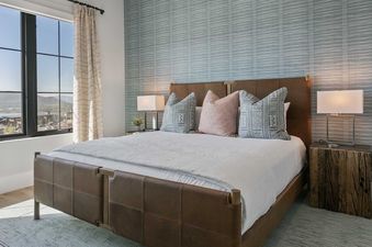 A serene bedroom at 11981 N Gemini Way SkyRidge featuring a brown leather bed, white duvet, patterned pillows, a textured blue accent wall, and large windows with mountain and water views.