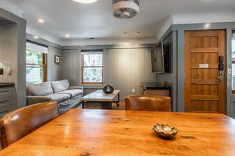 The living and dining area at Cedar Crest Stellars Jay Cottage 4 features a wooden dining table, leather chairs, a sofa, and large windows with a winter view.