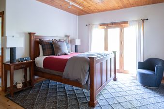 A cozy bedroom with a large wooden bed, nightstand, lamp, and a blue armchair, featuring warm wood accents and bright natural light.