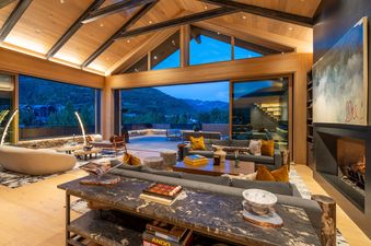 The luxurious living room of Opus Estate features vaulted wooden ceilings, comfortable seating, a fireplace, and large glass doors opening to a scenic mountain view.